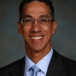 Smiling man with glasses wearing a suit and tie, set against a blurred background.