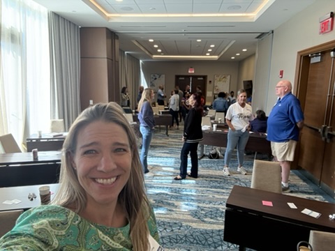 A woman smiling in the foreground, taking a selfie at a conference or workshop event, with people interacting and networking in the background. The setting features tables with name tags and a well-lit room.