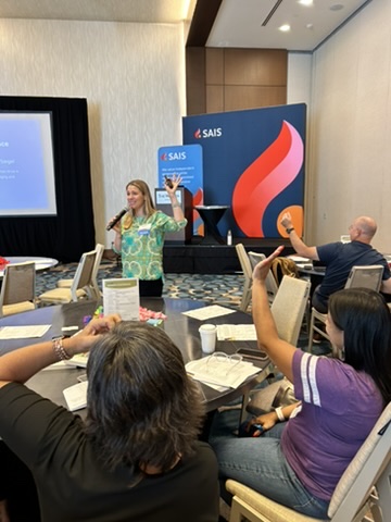 A woman speaking to an audience in a conference setting, with attendees raising their hands. There is a screen displaying information in the background and a brightly colored banner reading 'SAIS'.